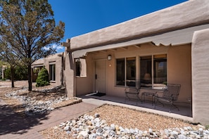A building with a beige exterior, a front door, and a patio area with two metal chairs and a table. - Artist Rd 7 | Cozy Fireplace, Pool Access, Centrally Located (Santa Fe)