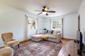 A living room with a beige sofa, a round coffee table, a beige armchair, a patterned rug, and a ceiling fan.