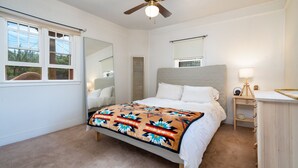 A bedroom with a bed, a ceiling fan, a bedside table with a lamp, a window with blinds, and a mirror. - Gomez Gardens | Fireplace, Yard, Private Patio (Santa Fe)