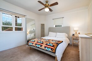 Quarto com cama, ventoinha no teto, mesa de cabeceira com abajur, janela com persianas e espelho.