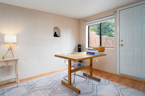 Quarto com uma mesa de madeira, uma luminária, uma pequena estante com livros e uma janela com vista para vegetação.