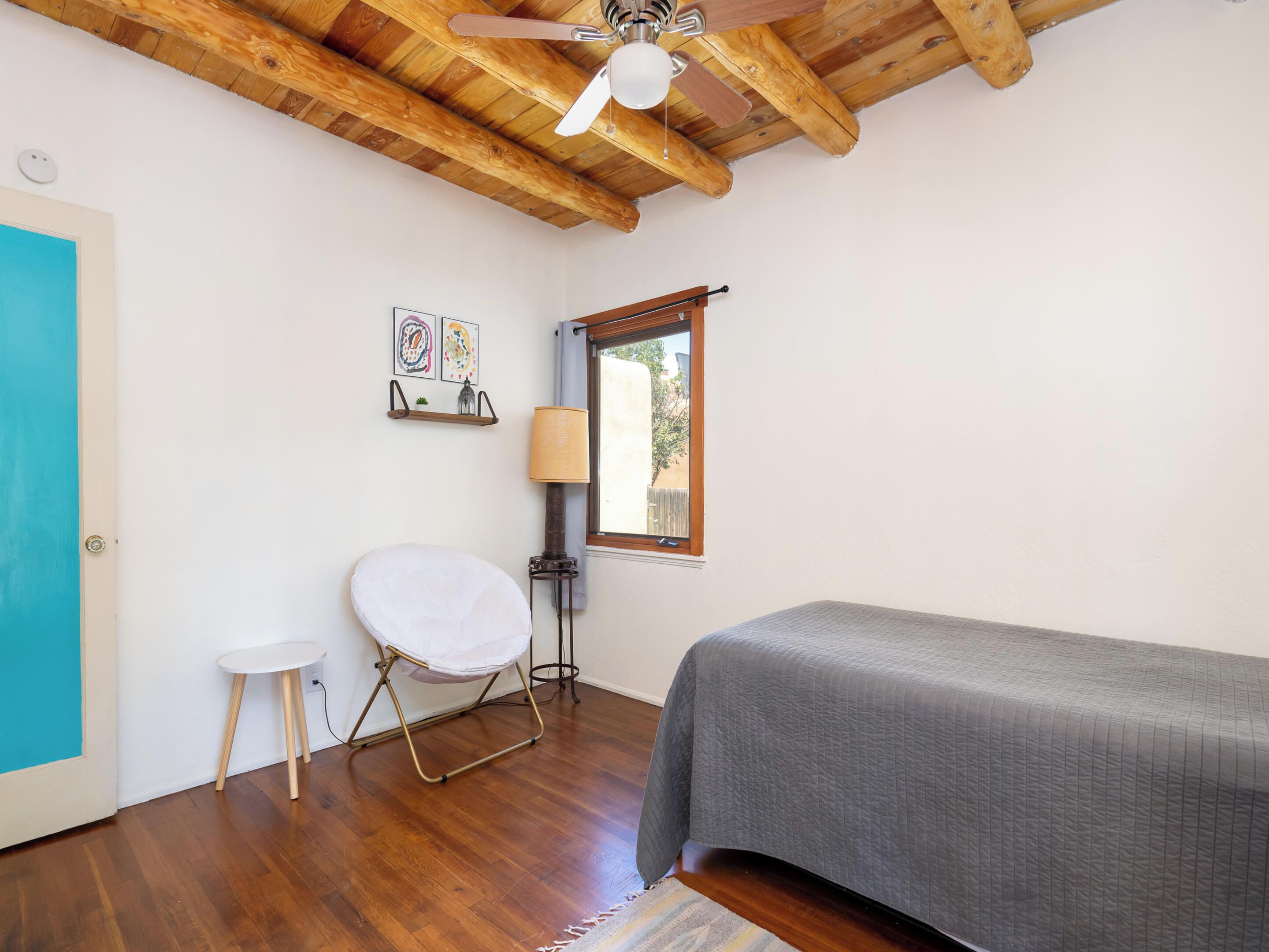 Un dormitori amb un llit, un tamboret petit, un ventilador de sostre, una finestra i una prestatgeria fixa a la paret amb obra d'art.