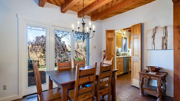 A dining area with a wooden table and chairs, a chandelier, and a view of the outside through large windows. - Puertas de Canyon Road | Patio, Fenced Yard! (Santa Fe)