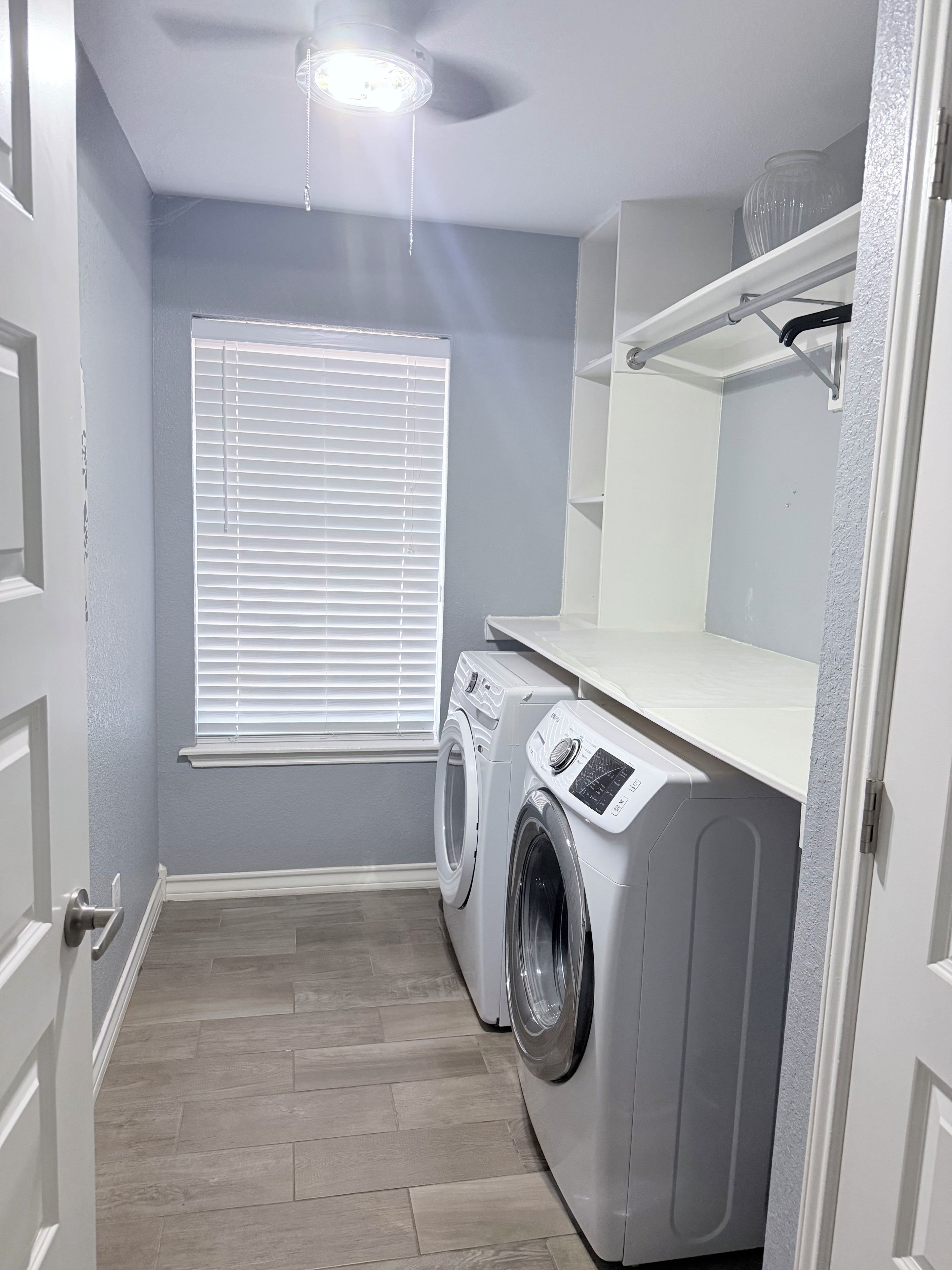 Une buanderie équipée d’un lave-linge, d’un sèche-linge, d’un ventilateur de plafond et d’une fenêtre avec des stores.