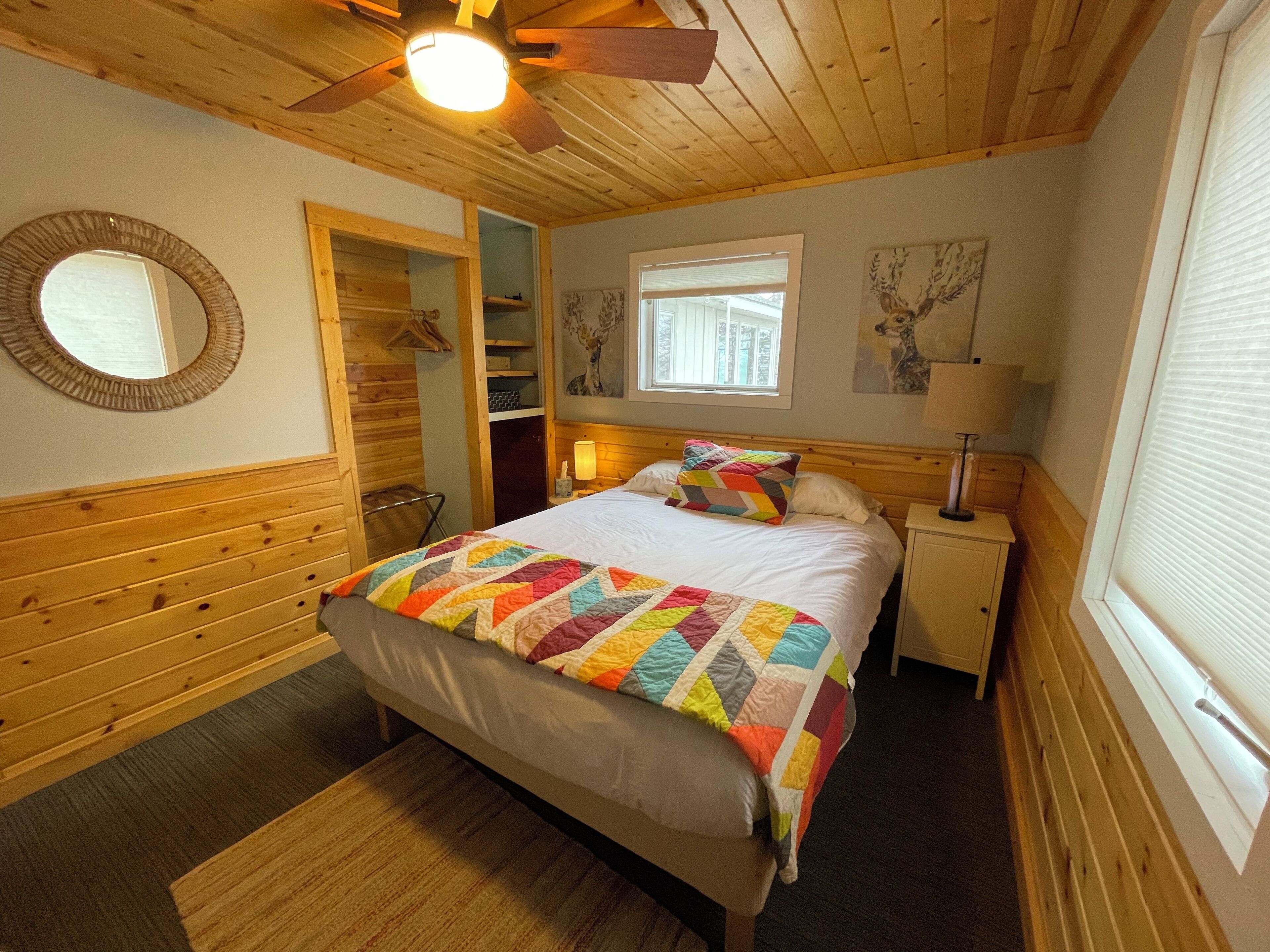 A cozy bedroom with a wooden ceiling fan, a bed with a colorful quilt, a nightstand with a lamp, and a window with blinds.