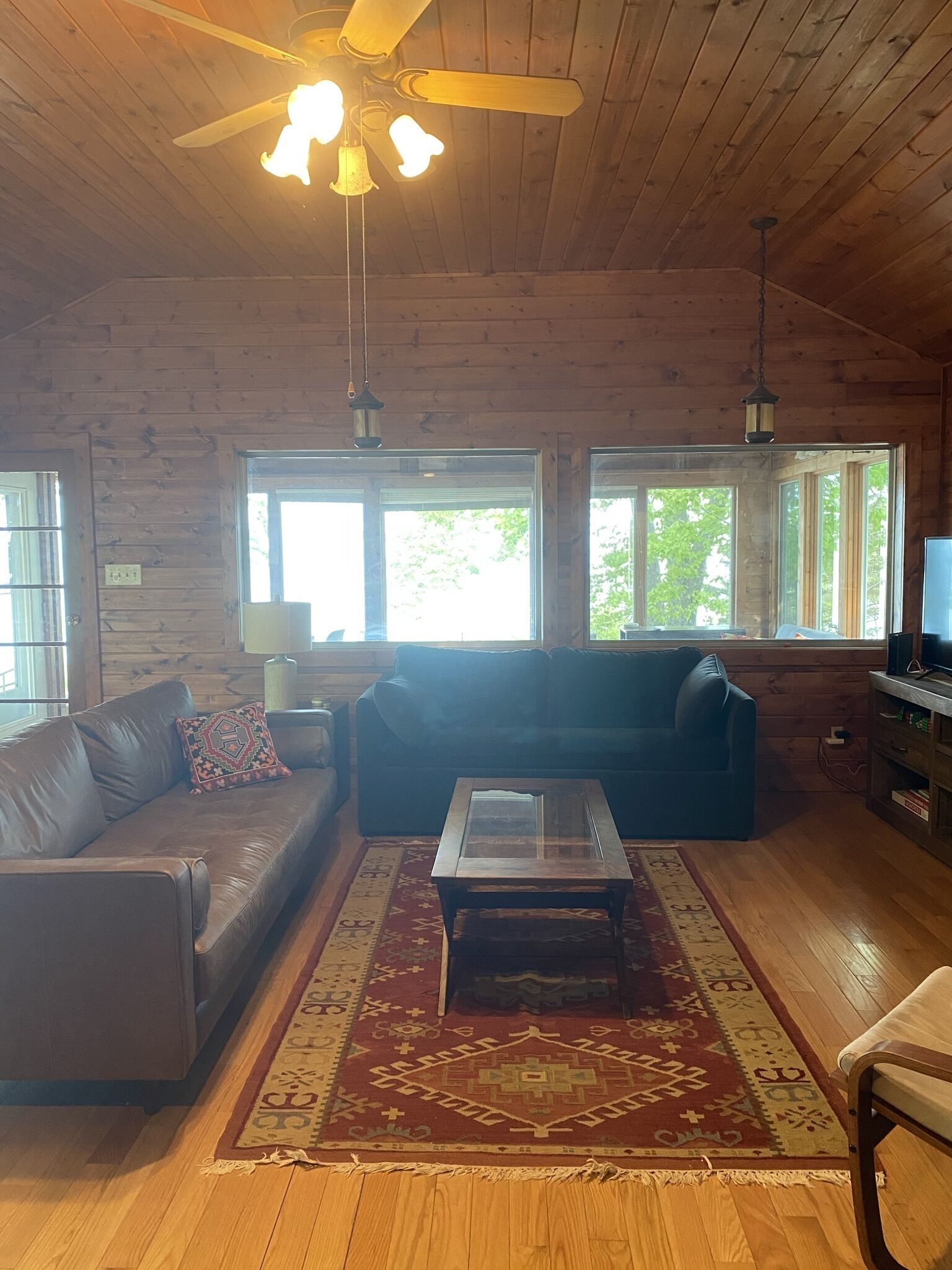 A cozy living room with a wooden ceiling, a ceiling fan, a leather sofa, a glass coffee table, and a TV stand.