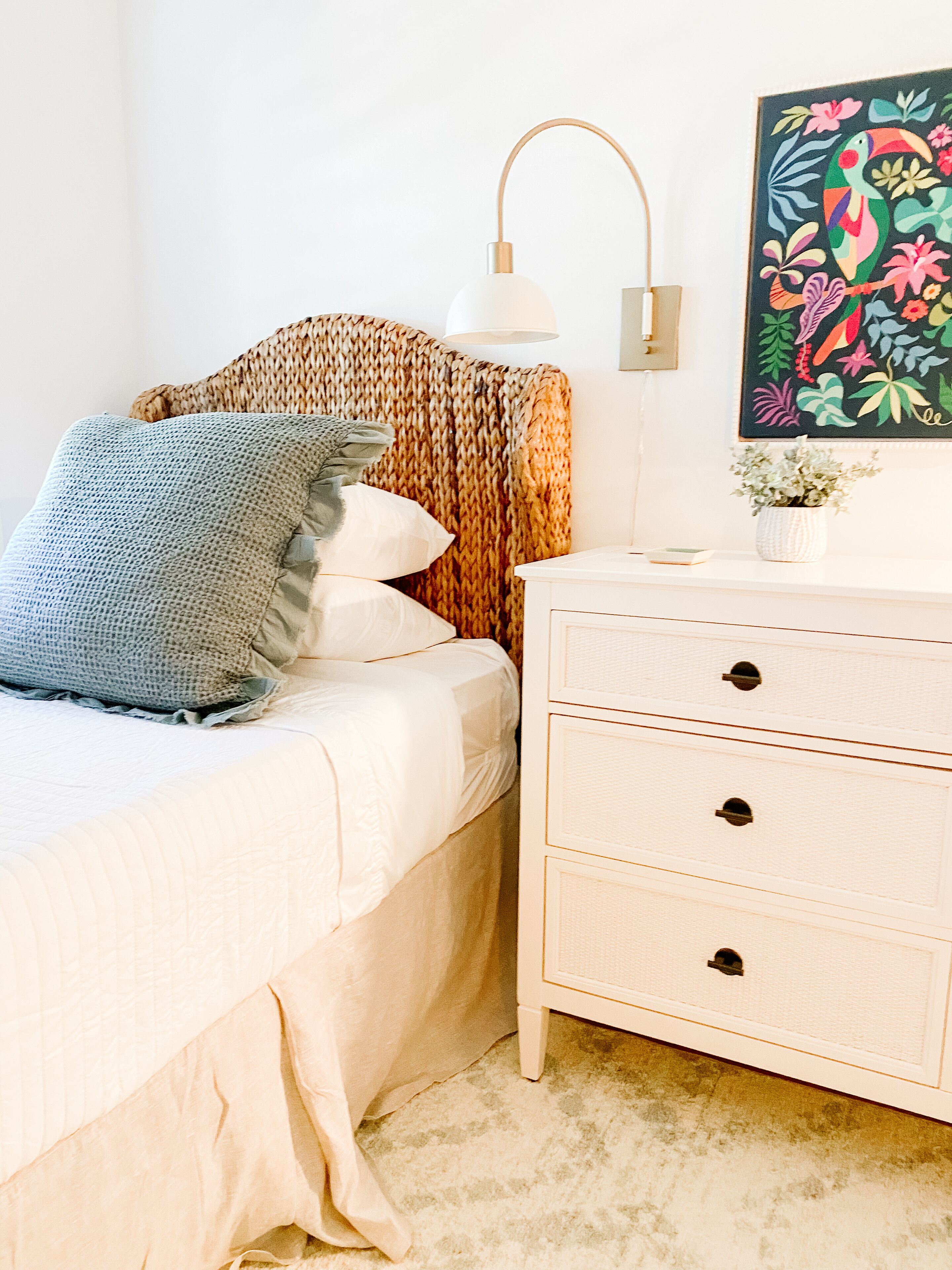 Un lit bien fait, avec une couette blanche et un oreiller bleu, une tête de lit en osier, une table de chevet blanche avec des tiroirs, une lampe de sol blanche et un tableau floral coloré accroché au mur.