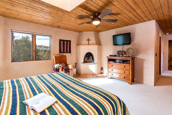 Un dormitorio con una cama grande, un ventilador de techo de madera, una chimenea, un televisor y una ventana con vistas a árboles.