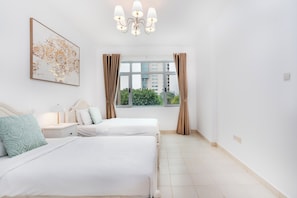 A hotel room with two beds, a large window with curtains, a chandelier, and a painting on the wall.