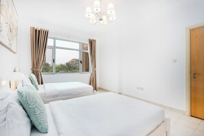 A hotel room with two beds, a chandelier, a window with curtains, and a door.