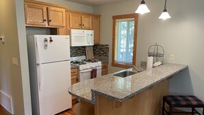 Una cocina con armarios de madera, refrigerador, estufa, microondas, encimeras de granito, un lavamanos y una ventana con persianas.