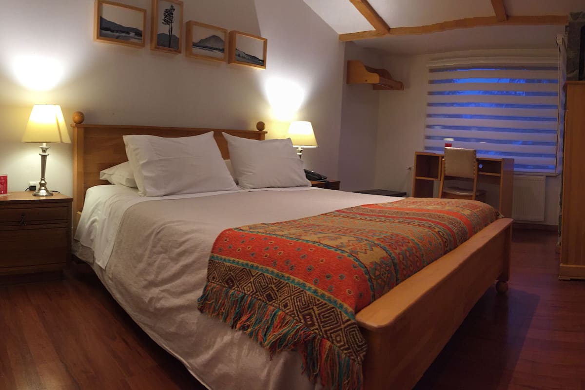 Quarto com cama de madeira, duas mesinhas de cabeceira com luminárias, uma escrivaninha pequena e uma janela com persianas.