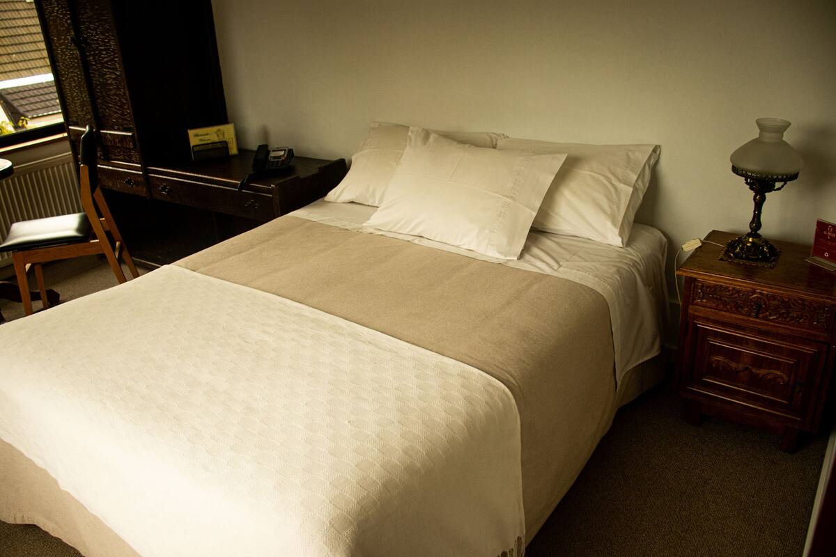 Uma cama bem arrumada com edredom branco e almofadas, uma mesa de cabeceira de madeira com abajur, e uma cadeira ao lado de uma escrivaninha com telefone.