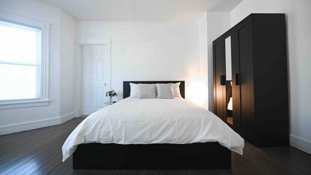 A modern bedroom with a large bed, a dark wardrobe, and a window with blinds. - FRESHLY RENOVATED 2 BR APARTMENT! (Ansonia)