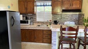 A kitchen with wooden cabinets, a refrigerator, a microwave, a sink, and a window with a view of greenery. - Villa Apartment in North Coast of Dominican Republic (Cabrera)