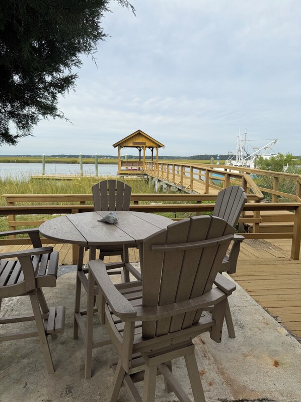 A wooden deck with a table and chairs, overlooking a body of water and a distant pier.