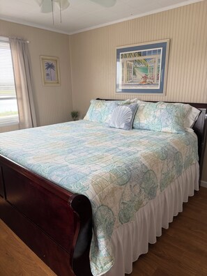 Un dormitorio con cama, ventana con cortinas, un cuadro en la pared y ventilador de techo.