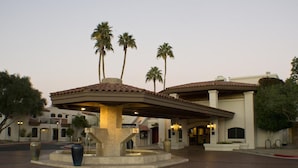 A hotel entrance with a covered patio, palm trees, and a fountain.