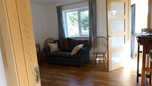 A room with a wooden floor, a dark sofa with cushions, a wooden chair, and a door with a frosted glass panel. - Delightful Bungalow Near Skibbereen & Baltimore (Skibbereen)