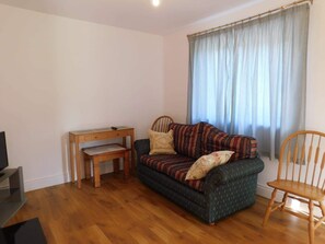 Ein kleines, möbliertes Zimmer mit einer Couch, einem Holzstuhl, einem Schreibtisch und einem Fernseher.