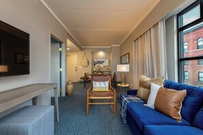 A hotel room with a blue sofa, a wooden chair, a flat-screen TV, a dining table with chairs, and a large window with curtains. - The Bradley Hotel (Fort Wayne)