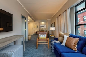 A hotel room with a blue sofa, a wooden chair, a flat-screen TV, a dining table with chairs, and a large window with curtains.