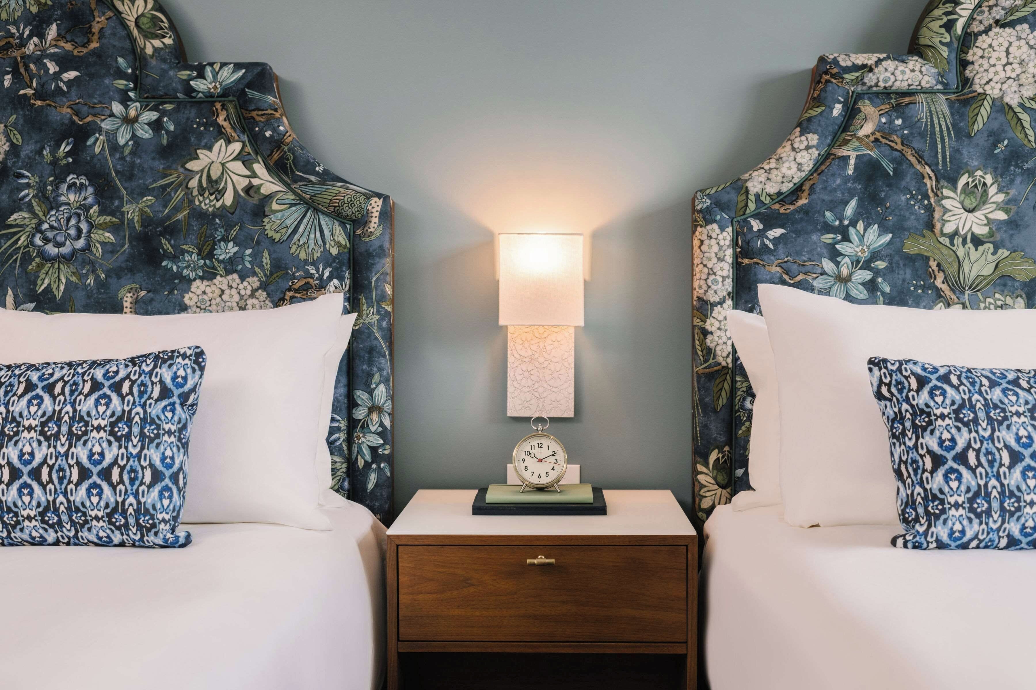 Una cama doble con cabecera floral y almohada a juego, una mesita de noche con lámpara y un pequeño reloj.