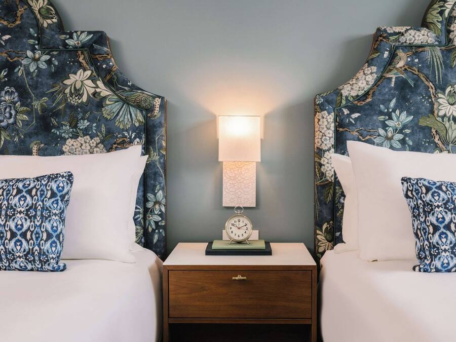Una cama doble con cabecera floral y almohada a juego, una mesita de noche con lámpara y un pequeño reloj.