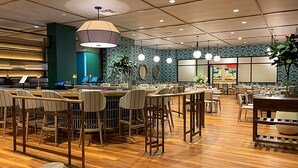 A restaurant with wooden flooring, multiple dining tables and chairs, hanging pendant lights, and a blue patterned wall. - The Bradley Hotel (Fort Wayne)