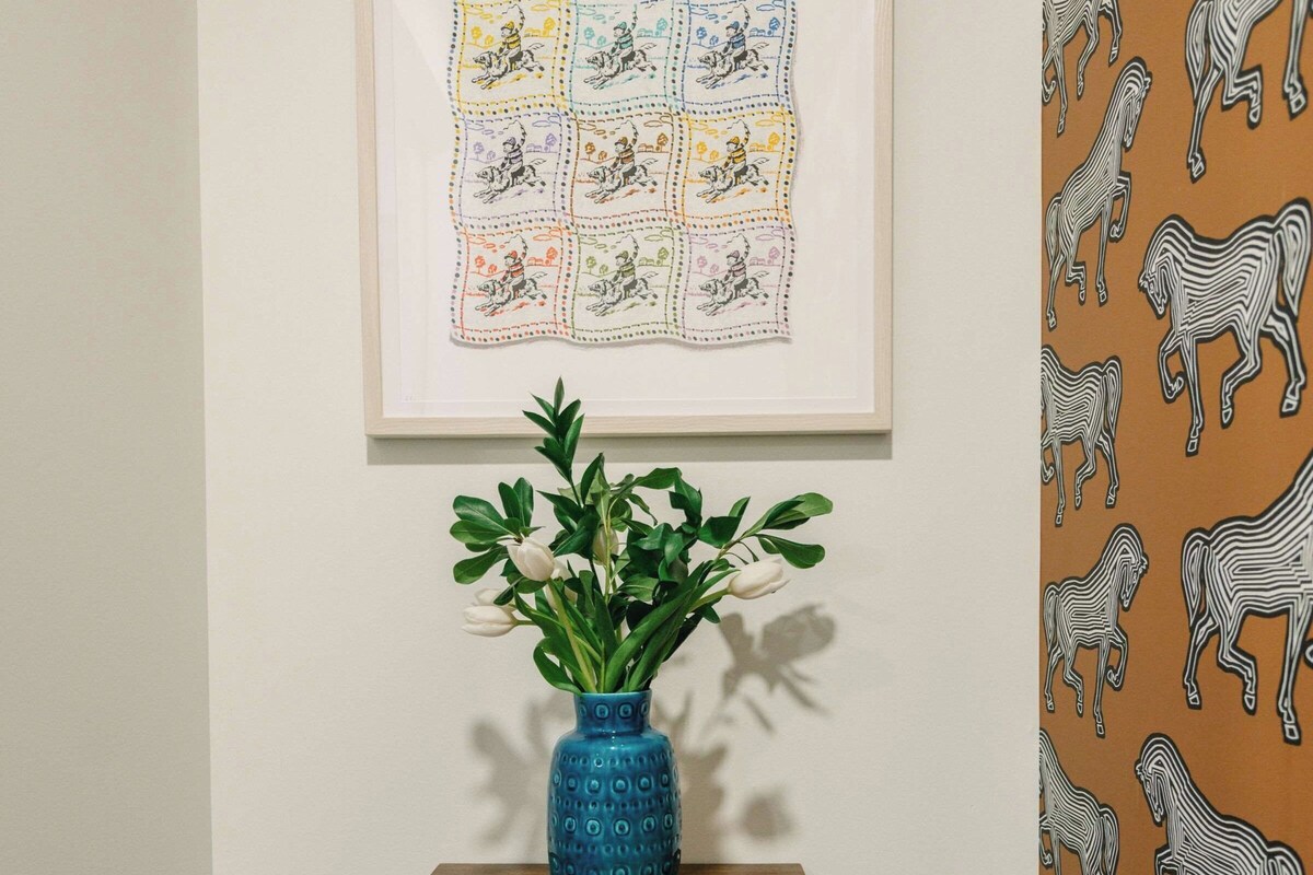 Una obra de arte enmarco en una pared, un jarrón azul con flores blancas sobre una mesita de madera, y un papel tapiz con motivos en la pared contigua.