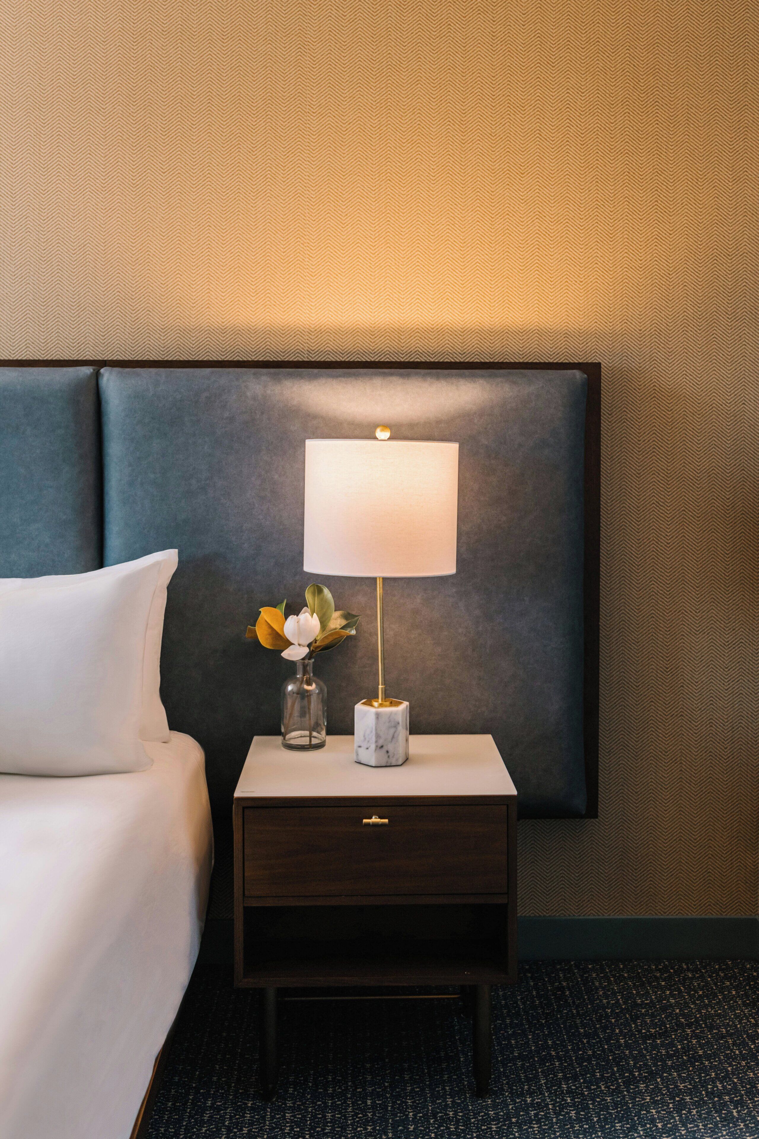 A hotel room with a bed, a nightstand with a lamp, a vase with flowers, and a marble-patterned object.