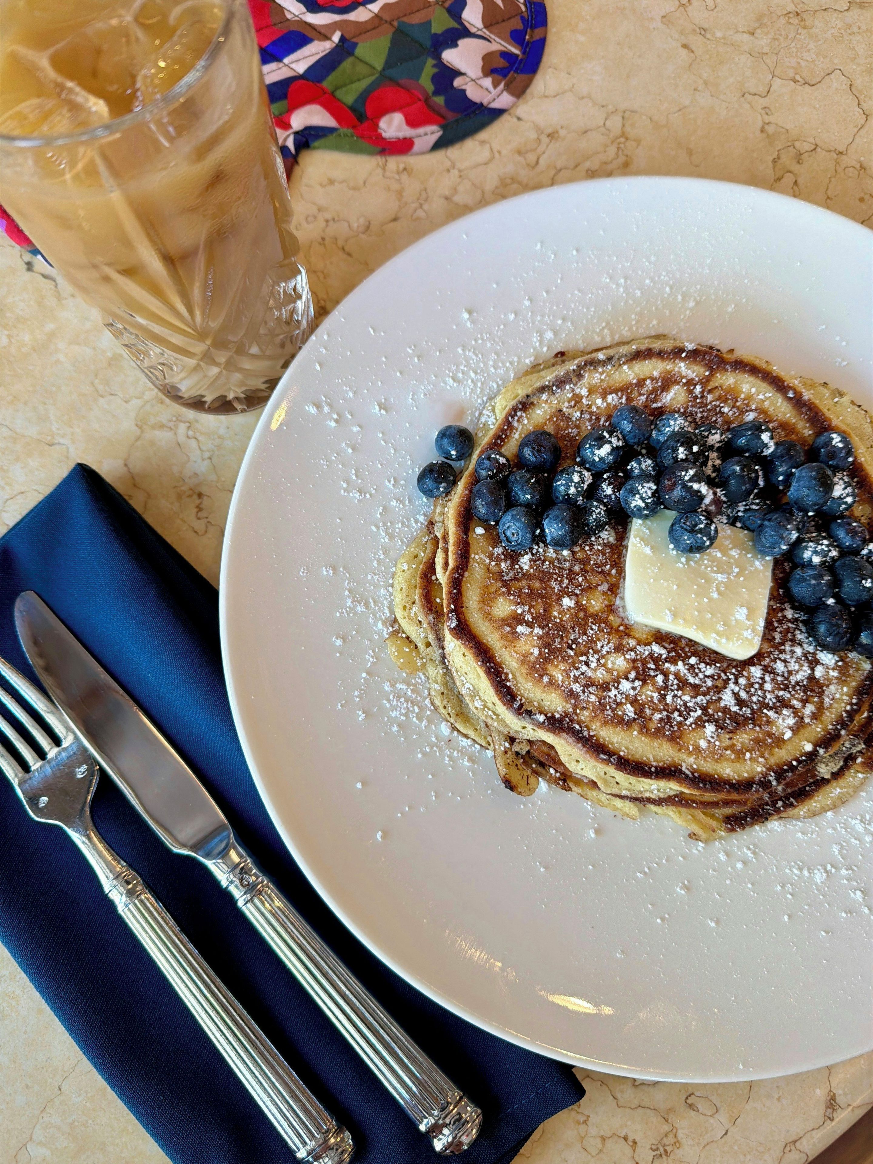 A plate of pancakes with blueberries and a pat of butter, accompanied by a glass of iced tea and cutlery on a table.