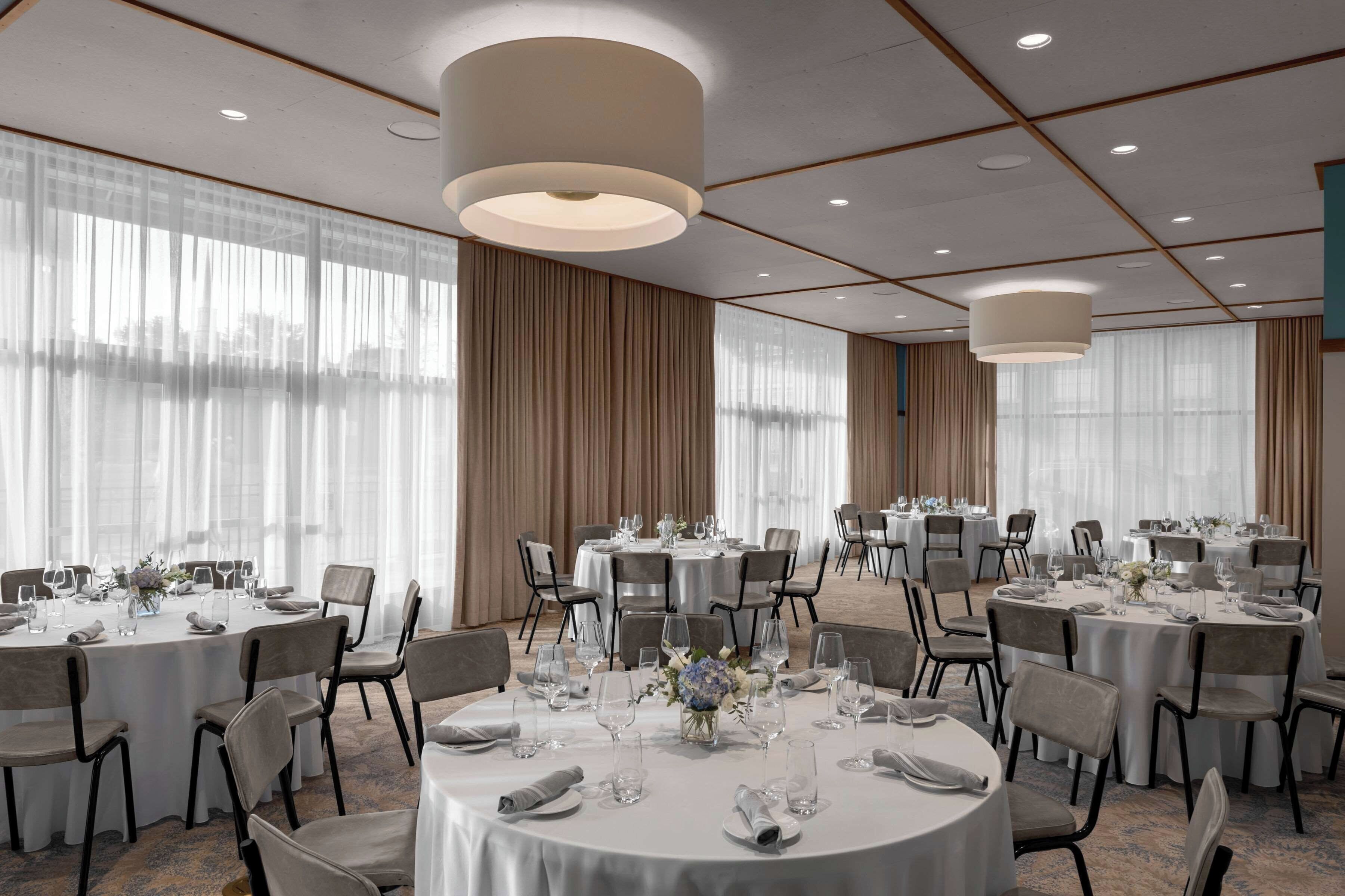 A hotel conference room with round tables, chairs, and large windows with curtains.