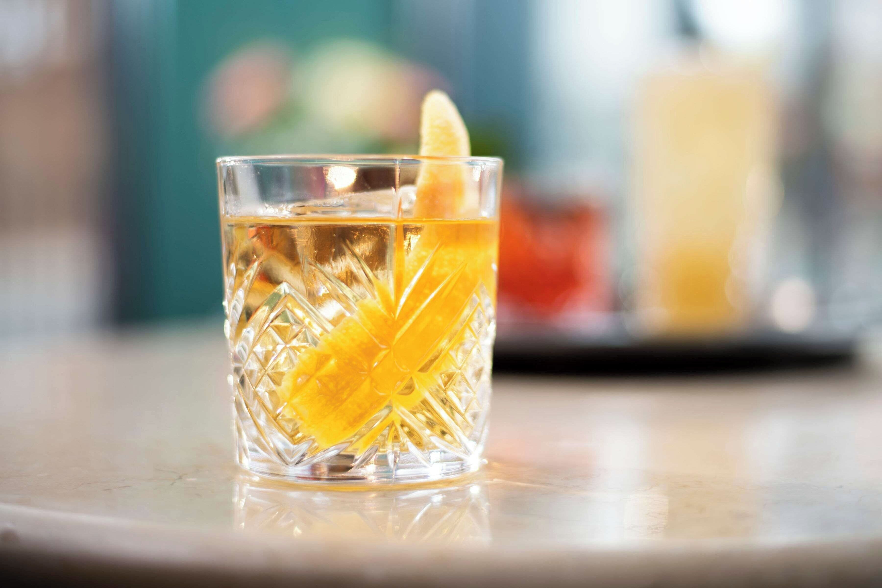 A clear glass with a textured pattern, containing a yellow liquid and a slice of orange, on a reflective surface.
