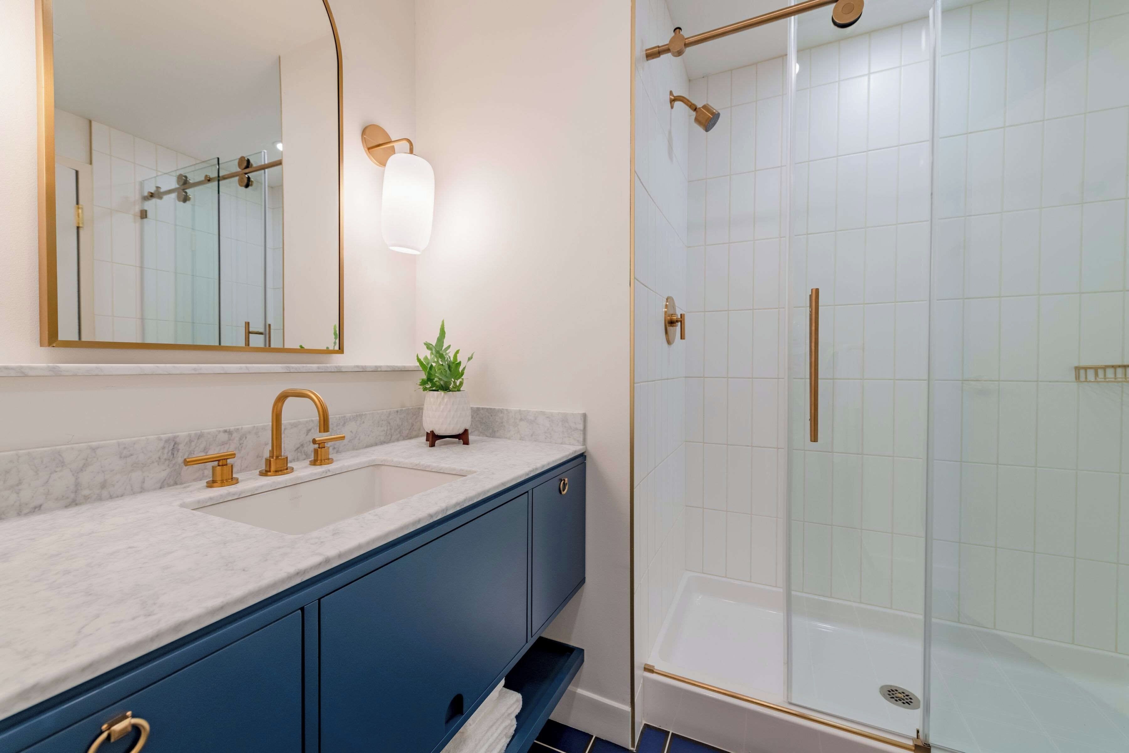 Un baño con encimera de mármol, grifo dorado, planta en maceta, cabina de ducha de cristal y un mueble azul.