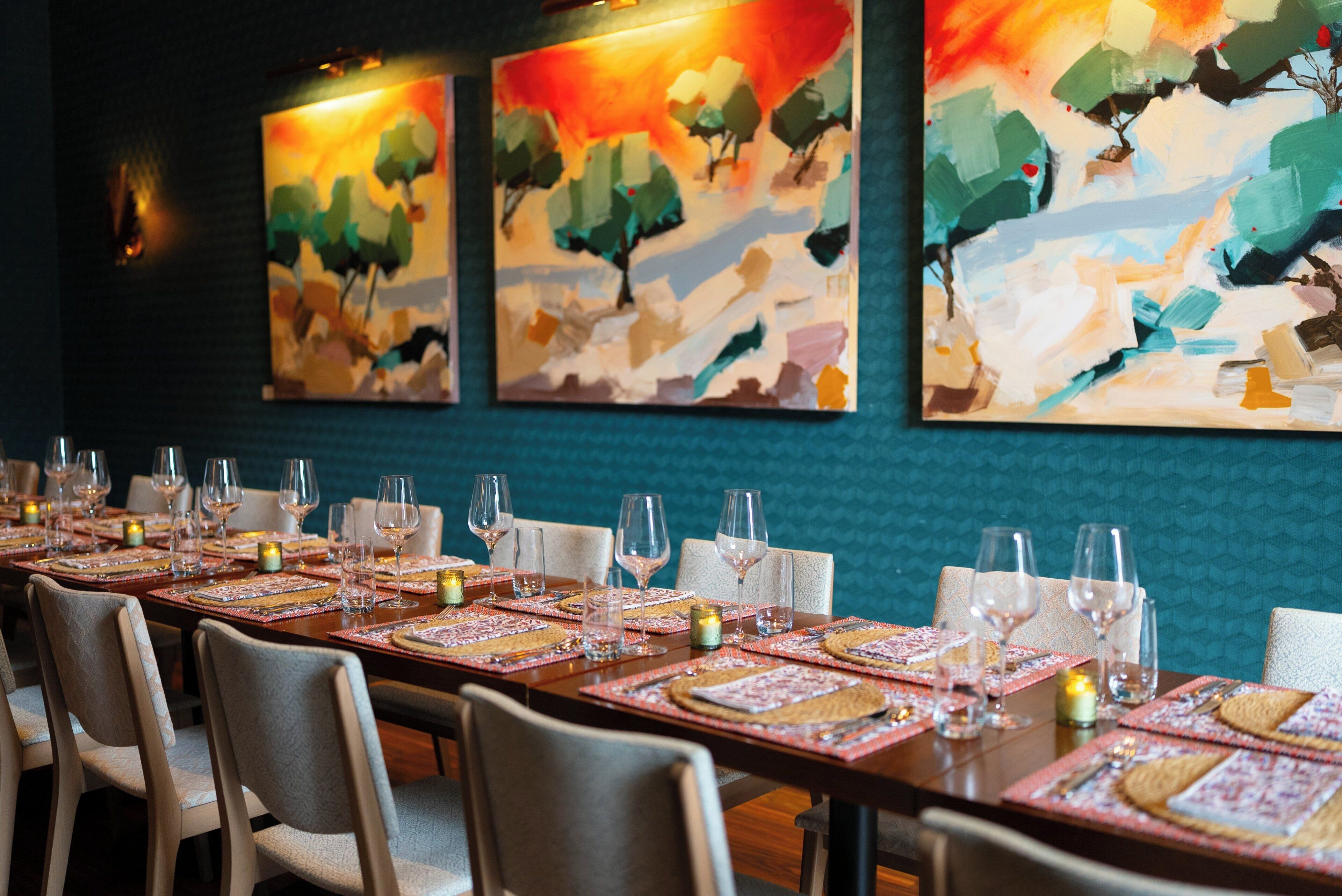 En restaurant med lange borde dækket til spisning, stole og kunst på væggen.
