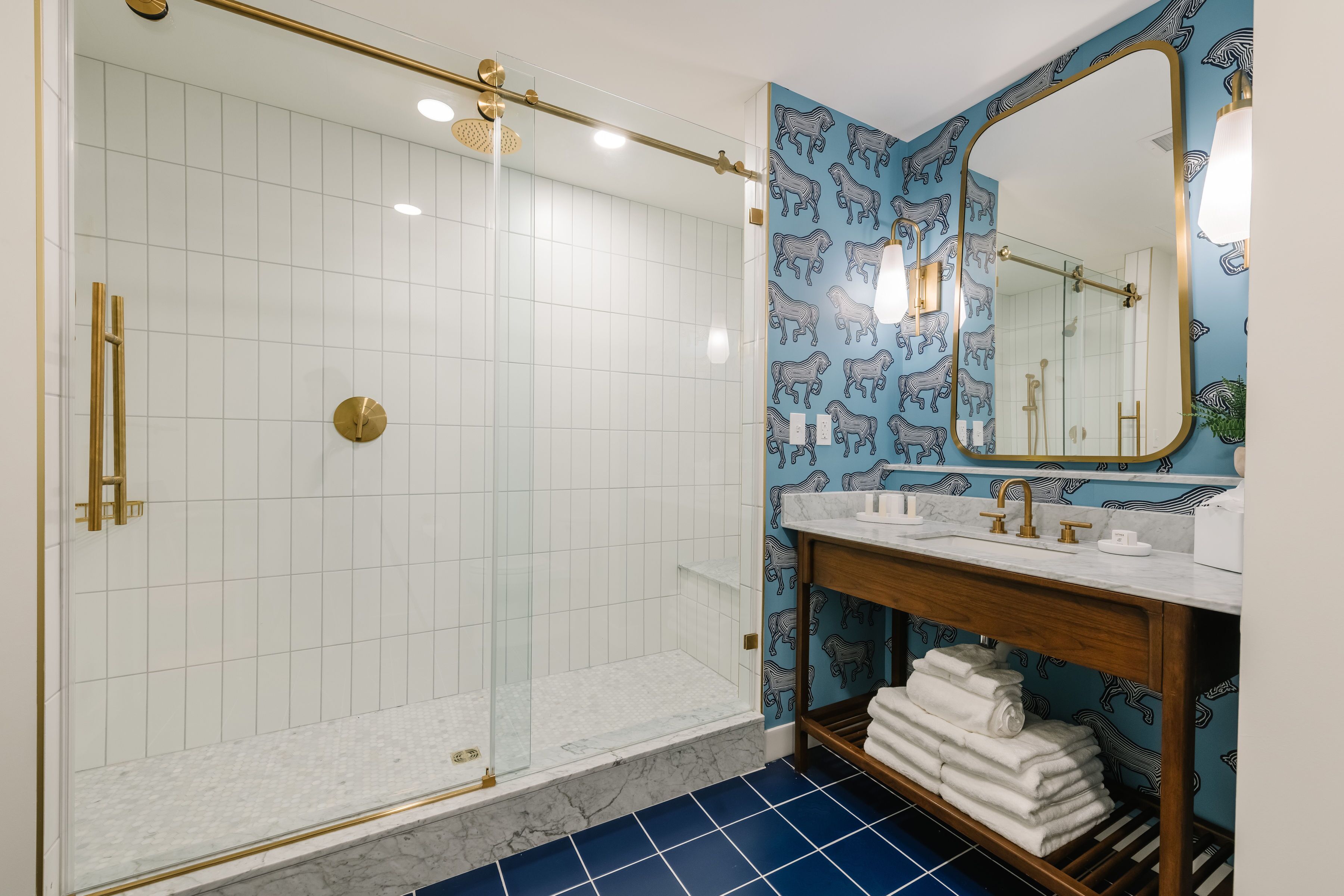 A bathroom with a glass-enclosed shower, white tiled walls, a blue patterned wall, a wooden vanity with a mirror, and a marble floor.