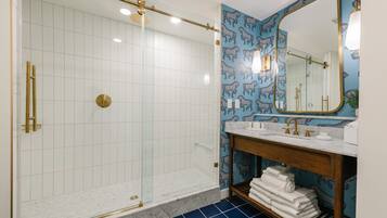 A bathroom with a glass-enclosed shower, white tiled walls, a blue patterned wall, a wooden vanity with a mirror, and a marble floor.