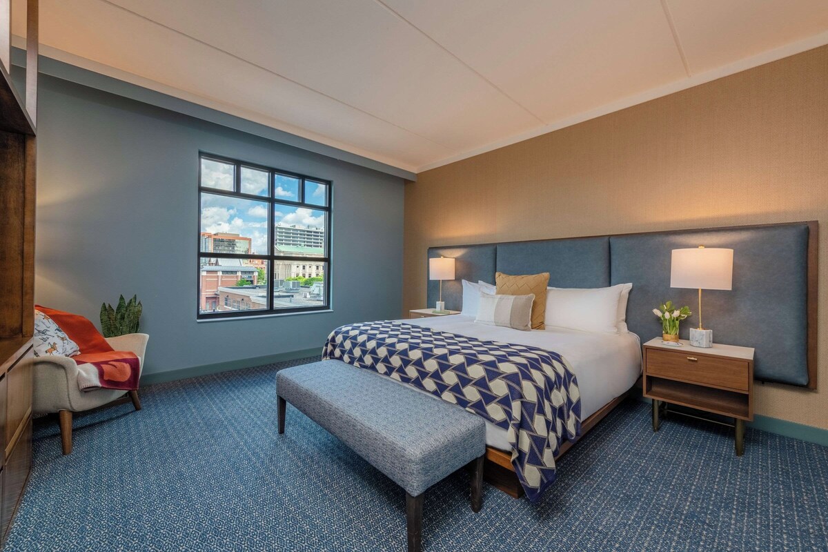A hotel room with a large bed, a bench, a nightstand, a lamp, and a window with a city view.