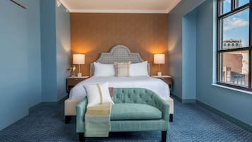 Een hotelkamer met een groot bed, twee bedlampen, een groene fauteuil en een raam met uitzicht op gebouwen.