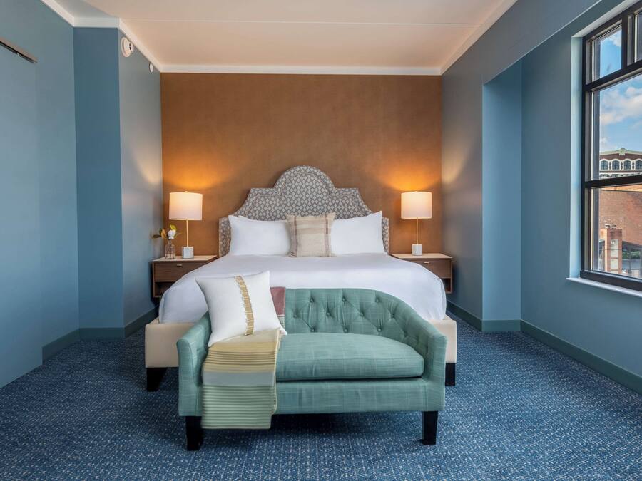 Een hotelkamer met een groot bed, twee bedlampen, een groene fauteuil en een raam met uitzicht op gebouwen.