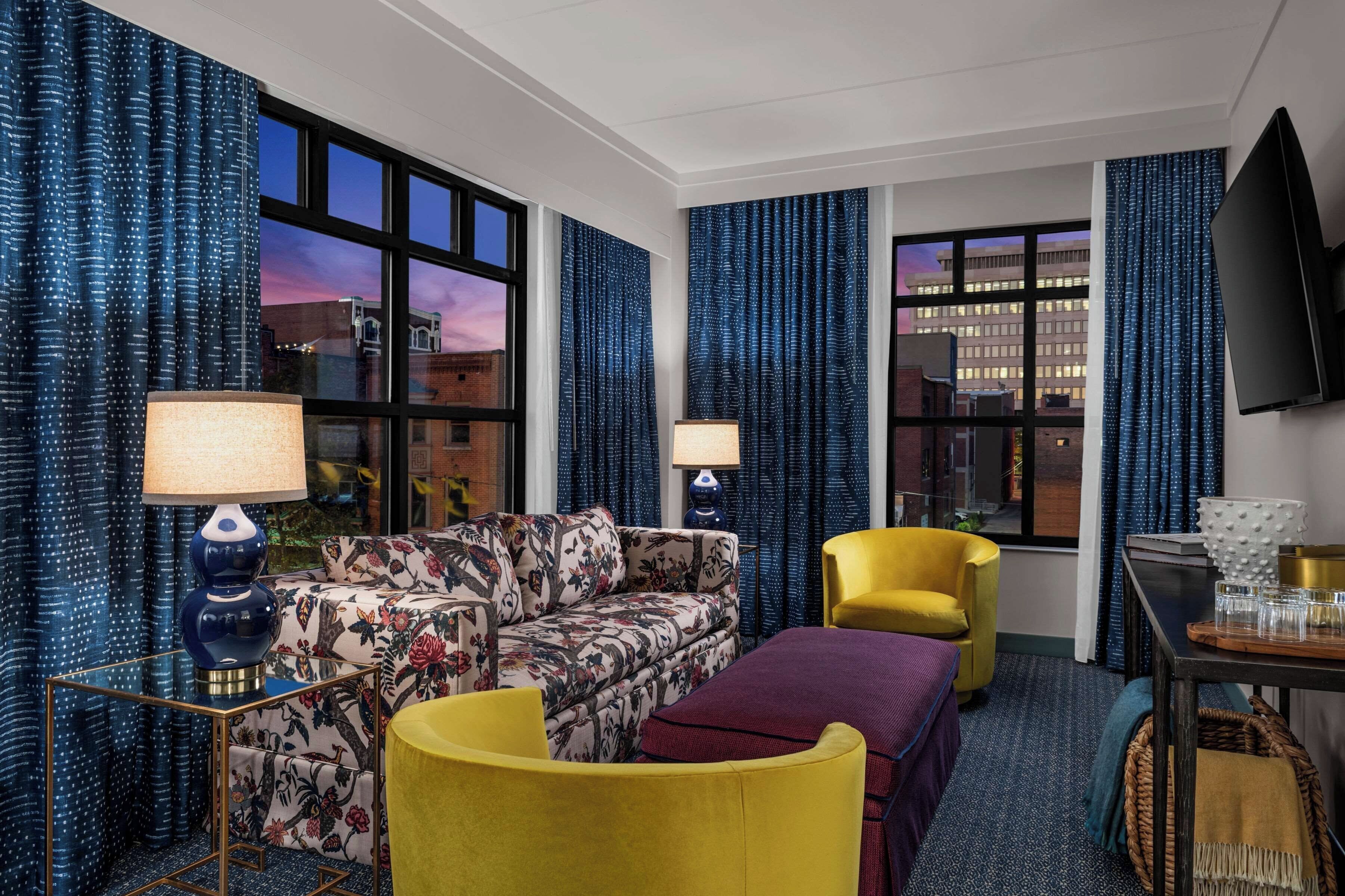 Ein Hotelzimmer mit einer Couch, einem Sessel, einem Hocker, einem Flachbildfernseher und einem Esstisch mit Gläsern und einer Schüssel.
