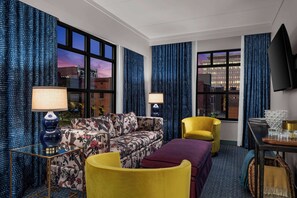 A hotel room with a sofa, armchair, and ottoman, a flat-screen TV, and a dining table with glasses and a bowl. - The Bradley Hotel (Fort Wayne)