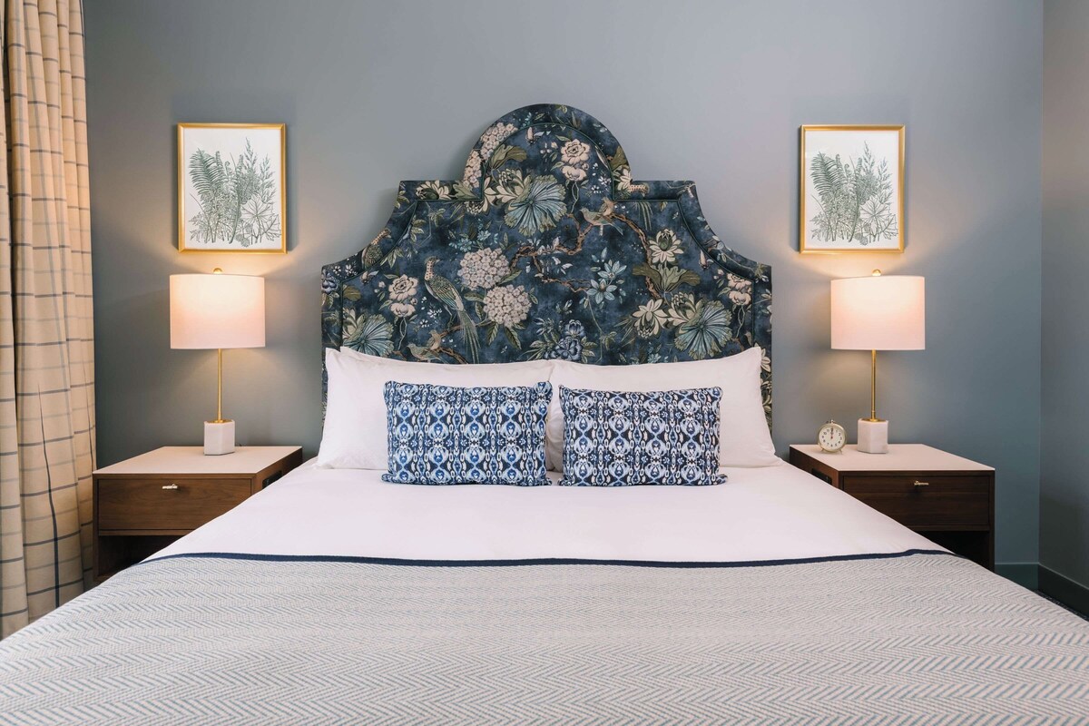 Una cama con cabecera floral, dos lámparas de noche e ilustraciones botánicas enmarcadas en la pared.