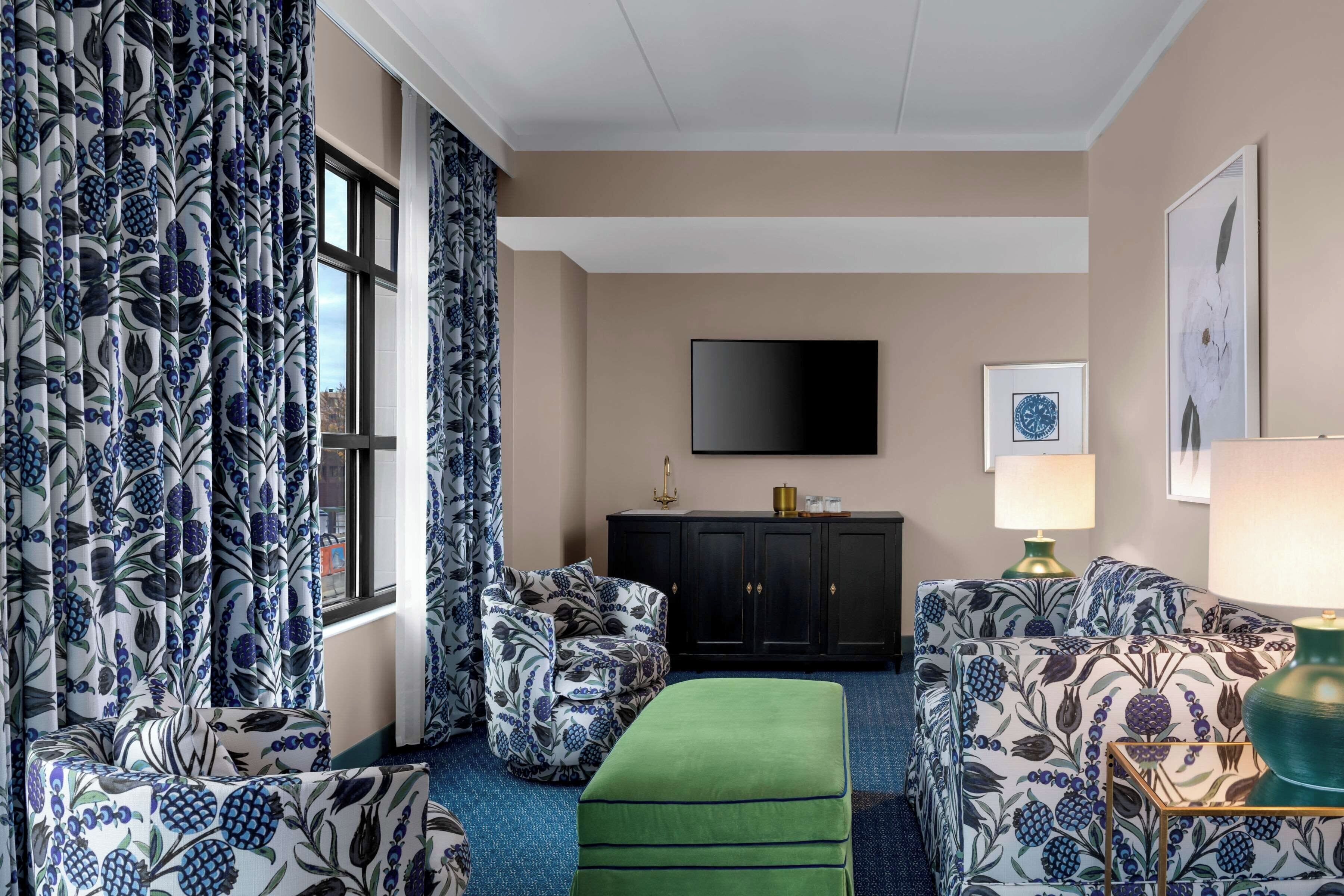 A hotel room with a large window, a flat-screen TV mounted on the wall, a green ottoman, and patterned furniture.