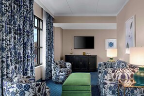 Une chambre d’hôtel dotée d’une grande fenêtre, d’un téléviseur à écran plat fixé au mur, d’un pouf vert et de meubles à motifs.