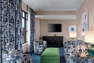 A hotel room with a large window, a flat-screen TV mounted on the wall, a green ottoman, and patterned furniture.