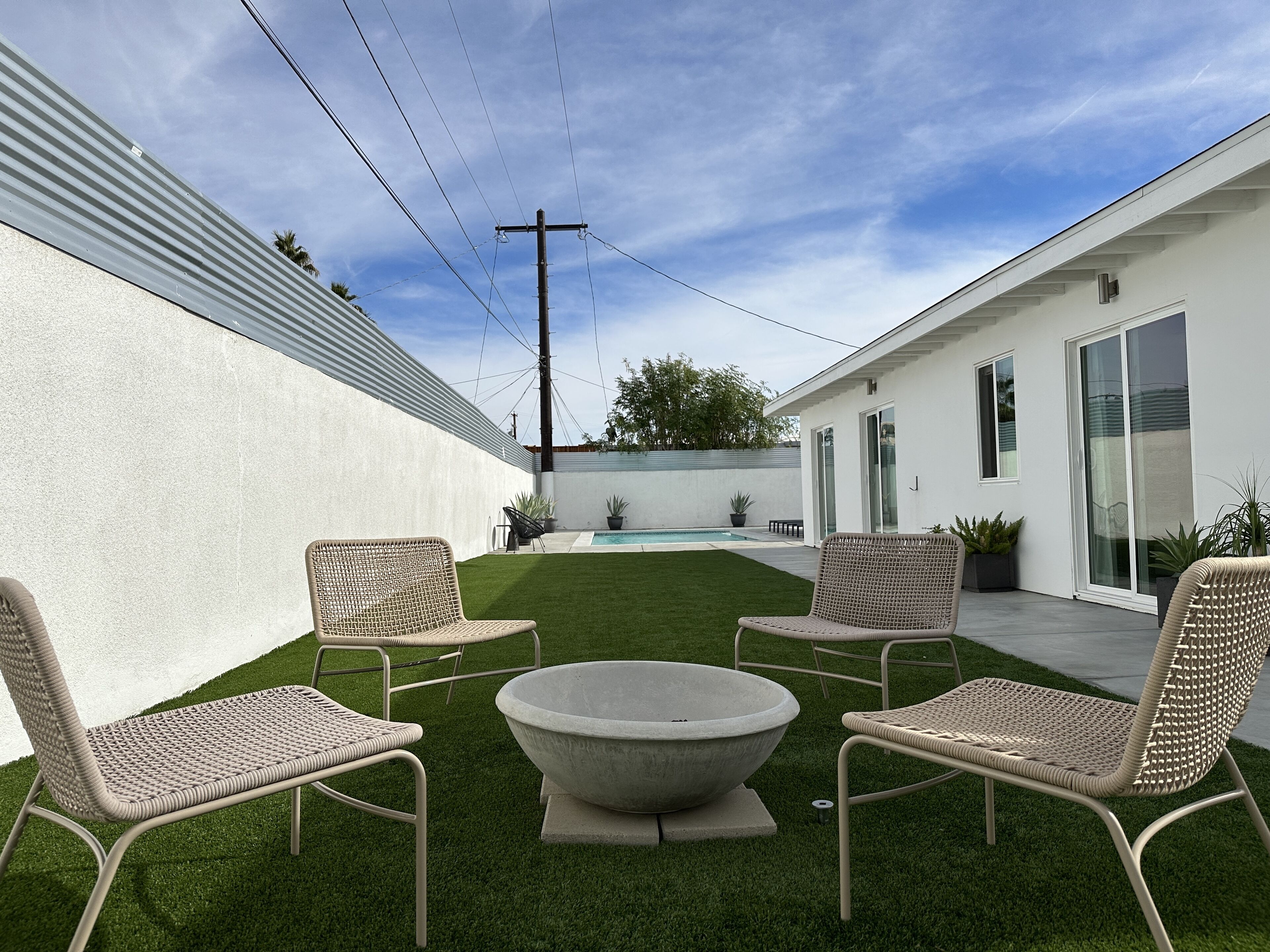 A modern outdoor patio with four wicker chairs, a concrete fire pit, and a swimming pool.