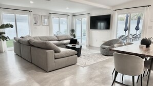 A modern living room with a beige sofa, a round grey chair, a dining table with chairs, and a flat-screen TV mounted on the wall. - Stylish Palm Springs House with Large Pool (Palm Springs)