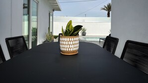 A black table with a patterned pot in the center, surrounded by black chairs, against a white wall with a glass door and a small pool in the background. - Stylish Palm Springs House with Large Pool (Palm Springs)